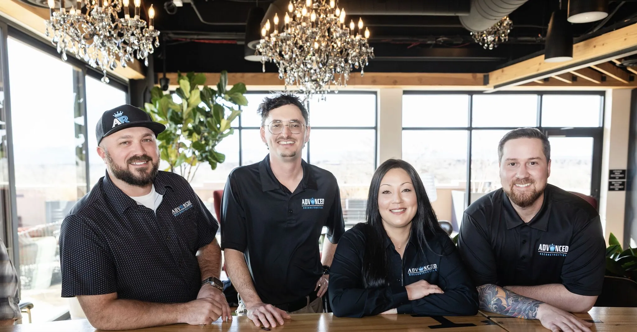 The Advanced Reconstruction team at Shift Workspaces, smiling together at a shared desk under chandeliers, showcasing their company culture and collaboration.